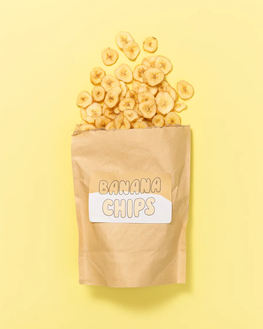 Vista de cima de um saco de papel kraft com o rótulo BANANA CHIPS e fatias de banana desidratada sobre fundo amarelo pastel.